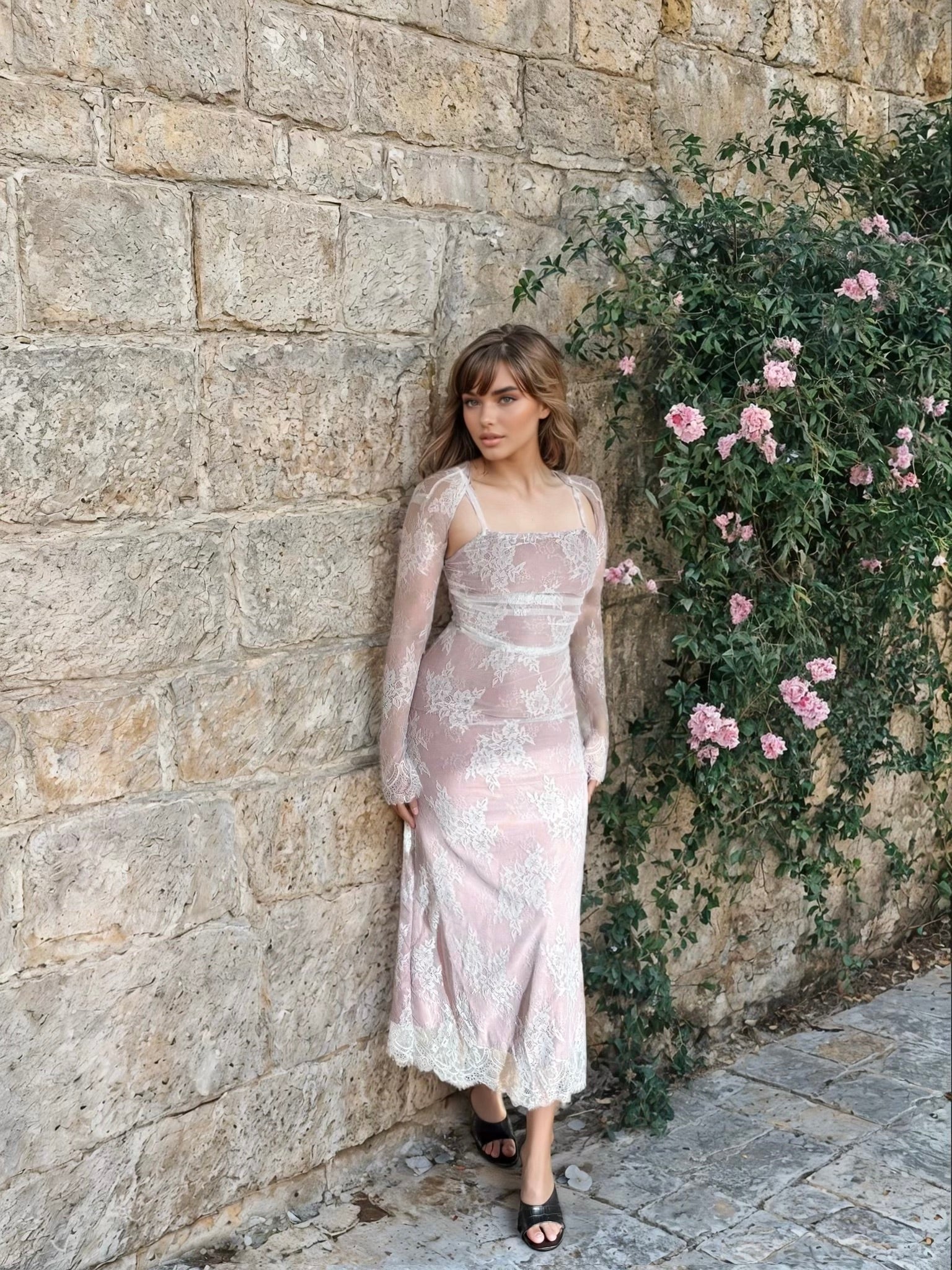 Woman in a lace dress standing against a stone wall with pink flowers.